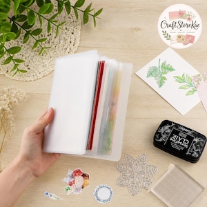 May include: A translucent plastic storage binder with multiple internal sleeves, containing various colorful paper inserts. Nearby are a black VersaFine CLAIR ink pad, a clear acrylic stamping block, a metal snowflake die, and floral stickers.