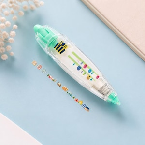 Puede incluir: Un dispensador de cinta correctora transparente, blanco y verde, con un dise&ntilde;o decorativo de casas. El dispensador est&aacute; sobre una superficie azul claro, con una tira de cinta decorativa con casas coloridas. Un cuaderno y flores est&aacute;n en el fondo.