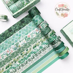 Puede incluir: Una colección de rollos de cinta decorativa washi en varios tonos de verde. Los patrones incluyen follaje botánico, escamas festoneadas, agujas de pino y motivos florales, junto con varios rollos de cinta adhesiva de color verde claro y oscuro.