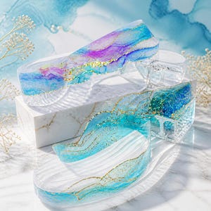 Puede incluir: Piezas decorativas de acrílico transparente con detalles en azul, morado y dorado. Las piezas tienen un diseño ondulado y se exhiben sobre una superficie de mármol blanco. El tema general es artístico y decorativo.