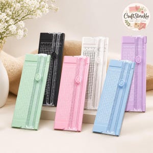 Puede incluir: Una colección de cinco cortadores de papel portátiles en colores menta pastel, negro, rosa pastel, blanco y azul pastel, cada uno con una guía de corte de plástico transparente, un mecanismo de cuchilla deslizante y una superficie cuadriculada.