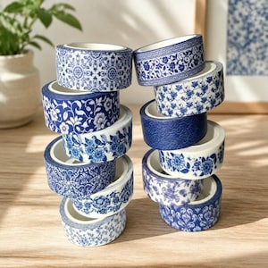 Puede incluir: Una pila de rollos de cinta adhesiva washi decorativa con diseños florales y geométricos en azul y blanco. Los rollos están apilados, mostrando una variedad de patrones. El fondo es una superficie de madera clara. Cada rollo mide aproximadamente 1,5 cm de alto.