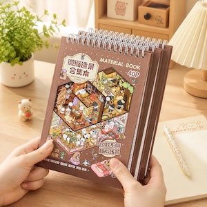 May include: A brown spiral-bound Material Book with a 3D miniature scene on the cover. The cover features a detailed illustration of a miniature room with cartoon characters. The book has the text "MATERIAL BOOK 40P" and other Chinese text.