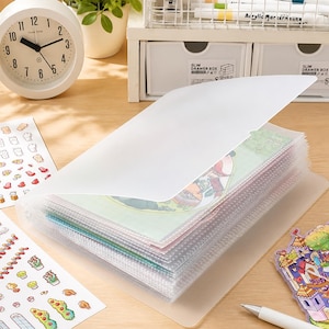 Puede incluir: Un álbum de fotos de plástico transparente con múltiples bolsillos, lleno de imágenes coloridas. El álbum está abierto, revelando el contenido. También se ven pegatinas y un reloj analógico blanco.