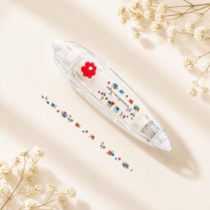 Puede incluir: Un dispensador de cinta correctora transparente con un acento floral rojo. El dispensador est&aacute; decorado con peque&ntilde;as flores azules, rojas y amarillas. La cinta en s&iacute; es una l&iacute;nea del mismo patr&oacute;n floral. El texto "Decoration Tape" est&aacute; impreso.