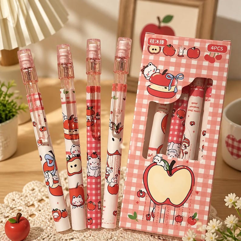 May include: Four pens with a white and pink gingham pattern featuring cartoon cats and red apples, accompanied by a matching rectangular cardboard box labeled 4PCS. The pens have clear plastic caps and clips, and the box displays apple graphics.