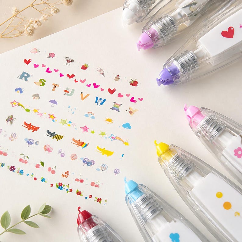 Puede incluir: Vista a&eacute;rea de varios dispensadores de cinta correctora transparente con acentos coloridos y dise&ntilde;os decorativos. Los dise&ntilde;os de la cinta incluyen corazones, estrellas y otras ilustraciones bonitas. Los dispensadores tienen acentos rosas, morados, amarillos, azules y rojos.