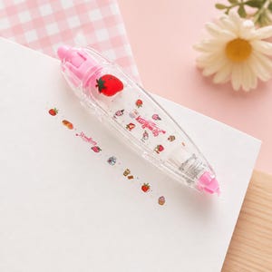 Puede incluir: Un dispensador de cinta correctora transparente, rosa y blanco con tem&aacute;tica de fresas. El dispensador tiene un gr&aacute;fico de fresa roja y las palabras "Strawberry" impresas en la cinta. La cinta est&aacute; decorada con ilustraciones de fresas y postres.