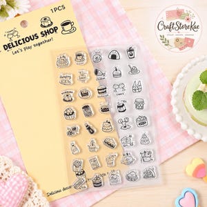 May include: A set of clear silicone stamps on a yellow backing card labeled DELICIOUS SHOP. The stamps feature black line art illustrations of various food and drink items including coffee, pudding, burgers, fries, and desserts.