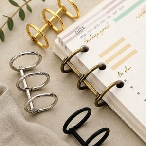 May include: A collection of metal loose-leaf binder rings in gold, silver, bronze, and matte black finishes. One bronze set is attached to a white planner page with the printed text DATE, MON, TUE, WED, THU, FRI, SAT, SUN, today goal, and to do.