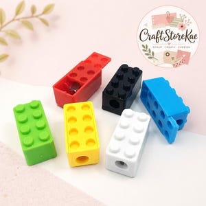 May include: A collection of six pencil sharpeners shaped like rectangular building blocks in red, black, blue, green, yellow, and white. Each sharpener features a top surface with eight circular studs and a front hole for inserting pencils.