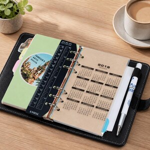 May include: A black leather-bound ring planner featuring a 2018 calendar on a translucent page, a black plastic ruler with white markings, a white pen with blue floral patterns, and a circular sticker depicting the Traveler's Hotel Seville.