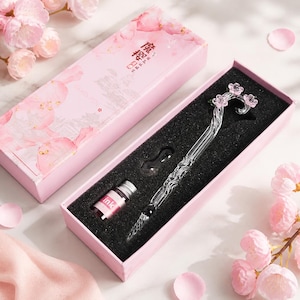 Könnte beinhalten: Ein transparenter Glas-Tauchfüller mit gedrehtem Schaft und einem dekorativen Ende, das mit drei kleinen rosa Blüten verziert ist, präsentiert in einer rosa rechteckigen Schachtel mit einer kleinen Flasche rosa Tinte und einer Glasablage.