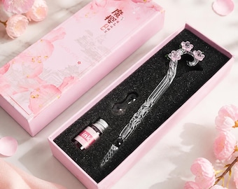 Op de afbeelding: Een transparante glazen dompelpen met een gedraaide steel en een decoratieve bovenkant met drie kleine roze bloesems, gepresenteerd in een roze rechthoekige doos met een bijpassend flesje roze inkt en een glazen pennenhouder in zwart schuim.