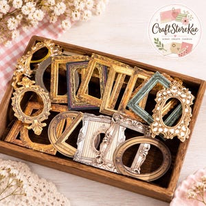May include: A collection of miniature decorative frames in various shapes, colors, and finishes, including gold, silver, and teal, arranged inside a rectangular wooden tray. The frames feature ornate, embossed, and textured details.