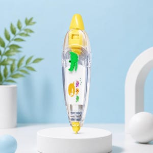 Puede incluir: Un dispensador de cinta correctora transparente y amarillo con un cocodrilo de dibujos animados verde y otros gr&aacute;ficos coloridos. El dispensador est&aacute; sobre una plataforma blanca sobre un fondo azul claro.