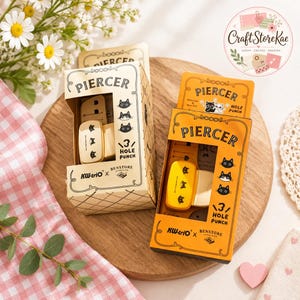 Può includere: Due perforatrici rettangolari in scatole individuali. Una scatola è color crema, l'altra è arancione. Entrambe le scatole riportano il testo PIERCER, 3 HOLE PUNCH, KW-triO e BENSTORE, insieme a illustrazioni di facce di gatto.