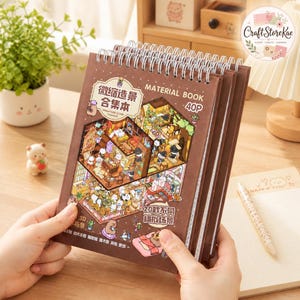 Könnte beinhalten: Ein braunes spiralgebundenes Materialbuch mit 40 Seiten voller Miniatur-Szenenaufkleber. Das Cover zeigt Illustrationen verschiedener Räume mit Zeichentrickfiguren, chinesischem Text und dem Aufdruck MATERIAL BOOK 40P.