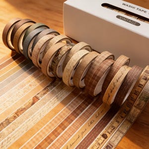 May include: A collection of vintage washi tape rolls in various shades of brown, beige, and tan. The rolls have different patterns, including script, plaid, and ruler designs. The box reads "Washi Tape Vintage".