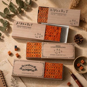 May include: A set of wooden rubber stamps in light brown sliding boxes. Each box contains orange rubber blocks with letters, numbers, and symbols. The boxes feature the text "AlphaBeT" or "BASICS" and "RUBBER STAMP SET (70 pcs)" printed on the lids.