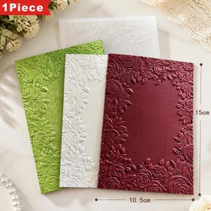 Puede incluir: Tres tarjetas rectangulares con bordes florales en relieve en verde, blanco y rojo oscuro. Cada tarjeta presenta un espacio central vacío rodeado de patrones de rosas realzados. Una medida indica que las tarjetas miden 15 cm por 10,5 cm.