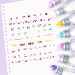 Puede incluir: Primer plano de una p&aacute;gina de cuaderno blanca con rollos de cinta decorativa. La cinta presenta ilustraciones coloridas de corazones, fresas, helados y otros dise&ntilde;os. Los dispensadores de cinta tienen cuerpos de pl&aacute;stico transparente y tapas de colores.
