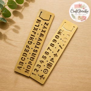 May include: Two rectangular gold-toned metal stencils featuring cut-out designs. One stencil displays the uppercase alphabet from A to Z, while the other features numbers 0 through 9, mathematical symbols, and various icons including a clock and arrow.