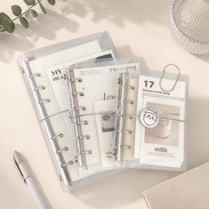 May include: Three clear, grid-patterned plastic ring binders of varying sizes, each containing white paper inserts with printed text and graphics. A silver elastic band wraps around the binders, featuring a small circular cat-themed charm.