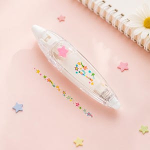Puede incluir: Un dispensador de cinta correctora blanco con una carcasa de pl&aacute;stico transparente. El dispensador tiene un adorno de estrella rosa y una tira de cinta decorativa con estrellas coloridas. Confeti en forma de estrella rosa, azul y amarillo sobre un fondo rosa.