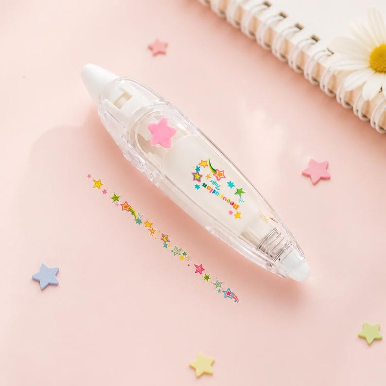 Puede incluir: Un dispensador de cinta correctora blanco con una carcasa de pl&aacute;stico transparente. El dispensador tiene un adorno de estrella rosa y una tira de cinta decorativa con estrellas coloridas. Confeti en forma de estrella rosa, azul y amarillo sobre un fondo rosa.
