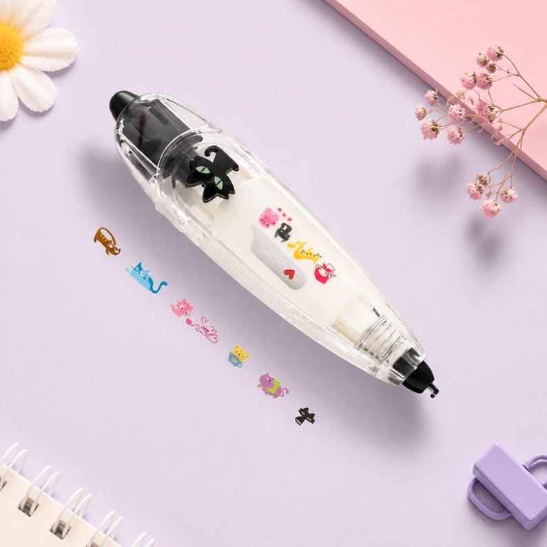 Puede incluir: Un dispensador de cinta correctora transparente, blanco y negro con un dise&ntilde;o de gato negro. El dispensador tiene una serie de pegatinas de gatos de colores. El fondo es una superficie de color p&uacute;rpura claro con una flor y un peque&ntilde;o objeto p&uacute;rpura.