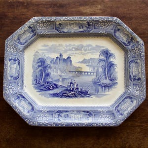 May include: An octagonal ceramic serving platter featuring a blue and white transferware pattern. The center depicts a landscape with a castle, a bridge, and figures near a river. The wide border displays floral motifs and architectural vignettes.