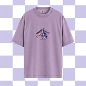May include: A lilac-colored t-shirt with a crew neck and short sleeves. The shirt features a colorful embroidered design of several stylized ribbons. The text "I can't have nice things" is printed below the design.