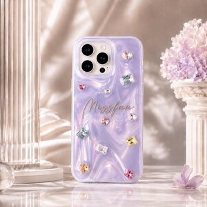 May include: A lavender phone case with a glossy, marbled texture featuring the gold script text "Missfan" and various multi-coloured, faceted crystal-like embellishments in heart, square, and rectangular shapes across the back surface.