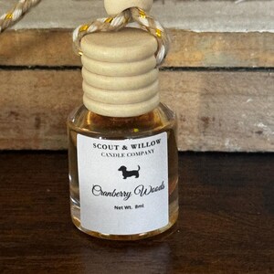 May include: A small glass bottle with a wooden cap and a decorative cord. The label reads "Scout & Willow Candle Company" and "Cranberry Woods" with a dachshund silhouette. Net weight is 8ml.