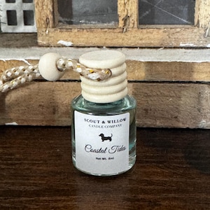May include: A small glass bottle with a wooden cap and a decorative string. The label reads "SCOUT & WILLOW CANDLE COMPANY" with a dachshund illustration and "Coastal Tides" text. Net weight is 8ml.
