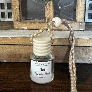 May include: A small, clear glass bottle with a wooden cap and a braided cord for hanging. The label reads "SCOUT & WILLOW CANDLE COMPANY" and "White Birch" with a dachshund silhouette. The cord is beige with gold accents.