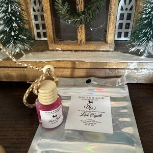 May include: A small, pink car air freshener with a wooden cap and a gold string. The label reads "Love Spell" and features a dog silhouette. A clear, iridescent bag and a white label with text are also visible.