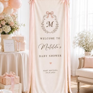 May include: A tall, cream-colored fabric banner hanging from a gold metal stand, featuring a pink bow, a floral wreath with the letter M, and the text: WELCOME TO Matilda's BABY SHOWER, a small heart, BABY MATILDA, and the date 09.18.2026.