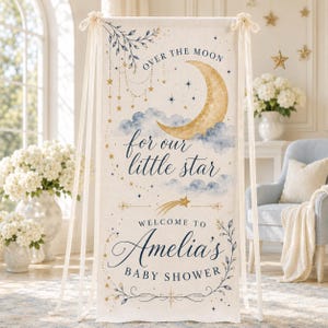 May include: A vertical fabric banner featuring a gold crescent moon, blue clouds, and gold stars. The text reads: OVER THE MOON for our little star WELCOME TO Amelia's BABY SHOWER. The banner is suspended by cream ribbons at the top corners.