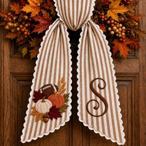 May include: A decorative fabric door hanger featuring brown and white vertical stripes with scalloped edges. One side displays an embroidered football, pumpkins, and autumn leaves. The other side features a large, dark brown embroidered letter S.