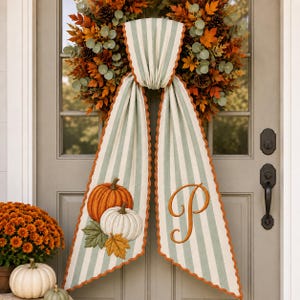 May include: A fall wreath featuring orange and brown leaves, pinecones, and eucalyptus, adorned with a long, light green and white striped fabric sash. The sash has scalloped edges, a pumpkin graphic on one side, and a large letter P on the other.