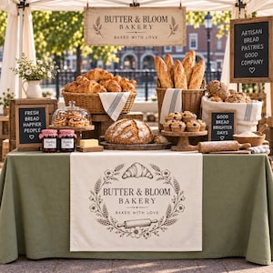 May include: A bakery stall display featuring a table with a green cloth, a runner with the text Butter & Bloom Bakery, baskets of croissants and baguettes, a large round loaf of bread, muffins, jars of jam, a rolling pin, and signs with text.