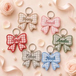 May include: Six embroidered bow keychains featuring a gingham pattern in beige, pink, red, green, gray, and blue. Each bow includes a silver metal keyring and is personalized with stitched names: Henry, Sarah, Sarah, Amelia, Ethan, and Noah.
