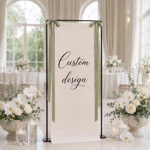 May include: A tall, rectangular, off-white fabric banner suspended from a black metal frame. The banner features the black script text Custom design and is adorned with two long, sage green satin ribbons tied at the top corners of the frame.