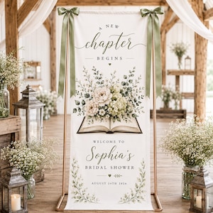 May include: A vertical white fabric banner suspended on a gold metal stand with sage green ribbons. The banner features the text A NEW CHAPTER BEGINS, an illustration of an open book with floral arrangements, and WELCOME TO Sophia's BRIDAL SHOWER.