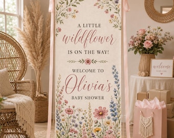 Könnte beinhalten: Ein vertikales Stoffbanner mit cremefarbenem Hintergrund und Illustrationen von Wildblumen oben und unten. Der Text lautet A LITTLE wildflower IS ON THE WAY! WELCOME TO Olivia's BABY SHOWER. Das Banner ist an hellrosa Bändern aufgehängt.