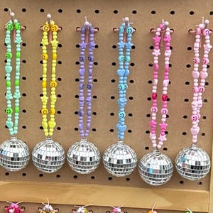 May include: A collection of fourteen hanging ornaments, each featuring a small silver mirrored disco ball attached to a beaded loop. The loops are constructed from various colorful plastic beads, including smiley faces, hearts, and stars in shades of pink, yellow, blue, green, and orange.
