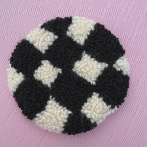 May include: A round, handmade coaster with a checkerboard pattern. The design features alternating black and cream-colored squares. The coaster is made of a textured, looped material, likely yarn or fabric, and is set against a pink background.
