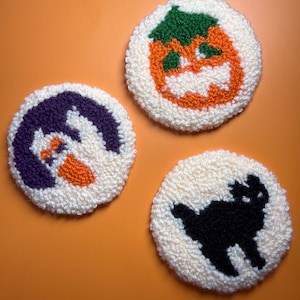 May include: Three circular punch needle coasters with a cream-coloured background. One coaster features an orange pumpkin with a green stem, another shows a purple ghost with orange accents, and the third displays a black cat silhouette.